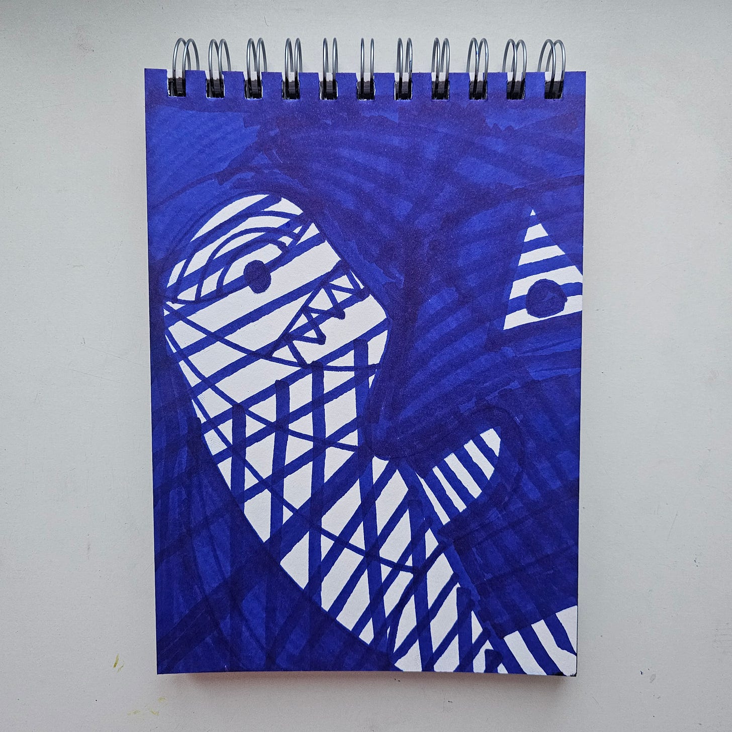 "A vertical marker drawing by artist FatbikeHero titled Mummy Boner. The artwork is done entirely in blue ink. It features a stylized, abstract figure resembling a wrapped mummy head with a grid-like pattern representing bandages. A single eye is visible on the left, and a triangular geometric shape floats on the right. The background is filled with heavy blue strokes."