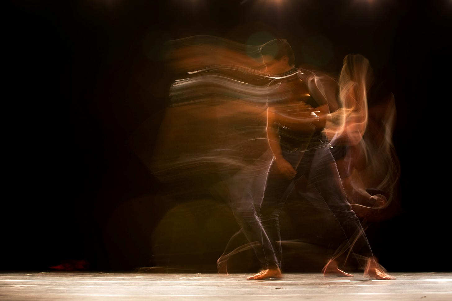 Two dancers in motion captured with long exposure photography, creating flowing light trails around their silhouettes as they move together on a wooden dance floor against a dark background. The ethereal streaks of light suggest graceful, continuous movement and intimate partnership in dance.