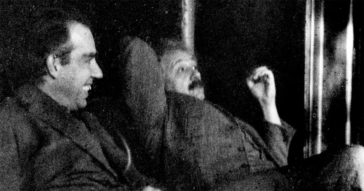 Fotografia histórica em preto e branco mostrando dois homens sentados lado a lado em um ambiente interno. Niels Bohr, à esquerda está de perfil, sorri amplamente e veste um terno escuro. Einstein, à direita aparece reclinado, com bigode, uma das mãos levantada acima da cabeça e expressão de riso, como se estivesse reagindo a uma conversa descontraída. A fotografia destaca a interação entre os dois.