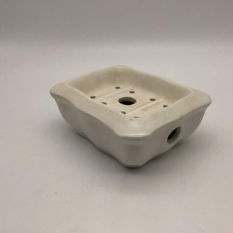 May include: A white ceramic soap dish with a rectangular shape and a raised rim. The dish has a drainage hole in the center and several smaller holes around the edges. The dish is designed to hold a bar of soap and allow water to drain away.