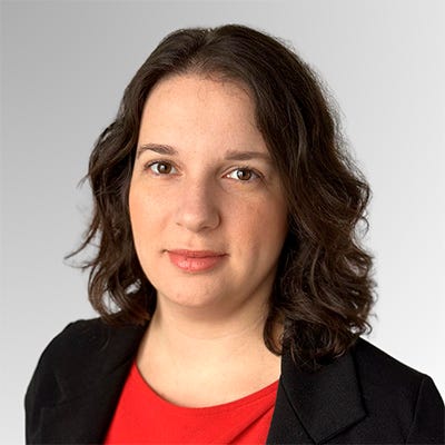Mulher de cabelos olhos castanhos, cabelos escuros e ondulados, sorri levemente e usa uma blusa vermelha sob blazer preto Mulher de cabelos olhos castanhos, cabelos escuros e ondulados, sorri levemente e usa uma blusa vermelha sob blazer preto