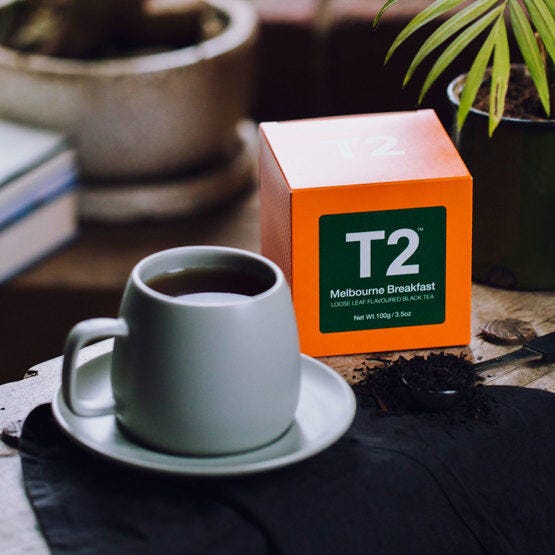 A picture of a grey cup and saucer filled with a dark liquid next to a square orange box with a dark green label on the front that says 'T2 Melbourne Breakfast', and in front is a teaspoon overflowing with loose leaf tea
