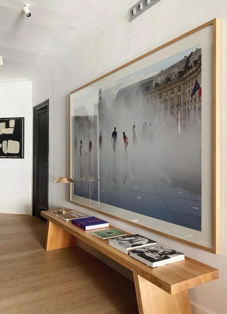 This may contain: an art work hangs on the wall above a wooden table with books and magazines under it