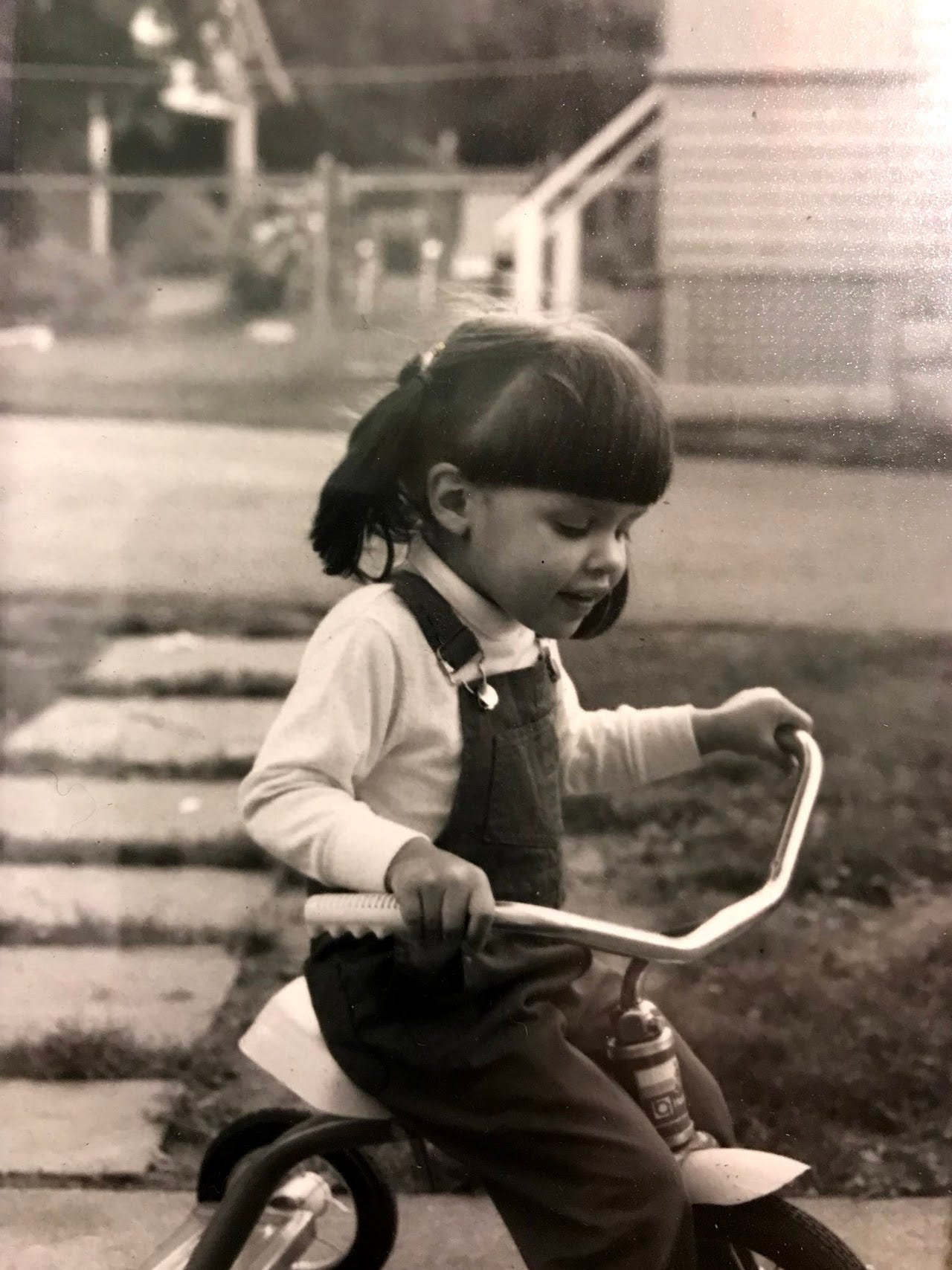 A black and white photo of a little girl in profile on a tricycle. In the background is a yard with a stone path and a house partially visible beyond it. The girl looks down at the handlebars with a slight smile on her face.