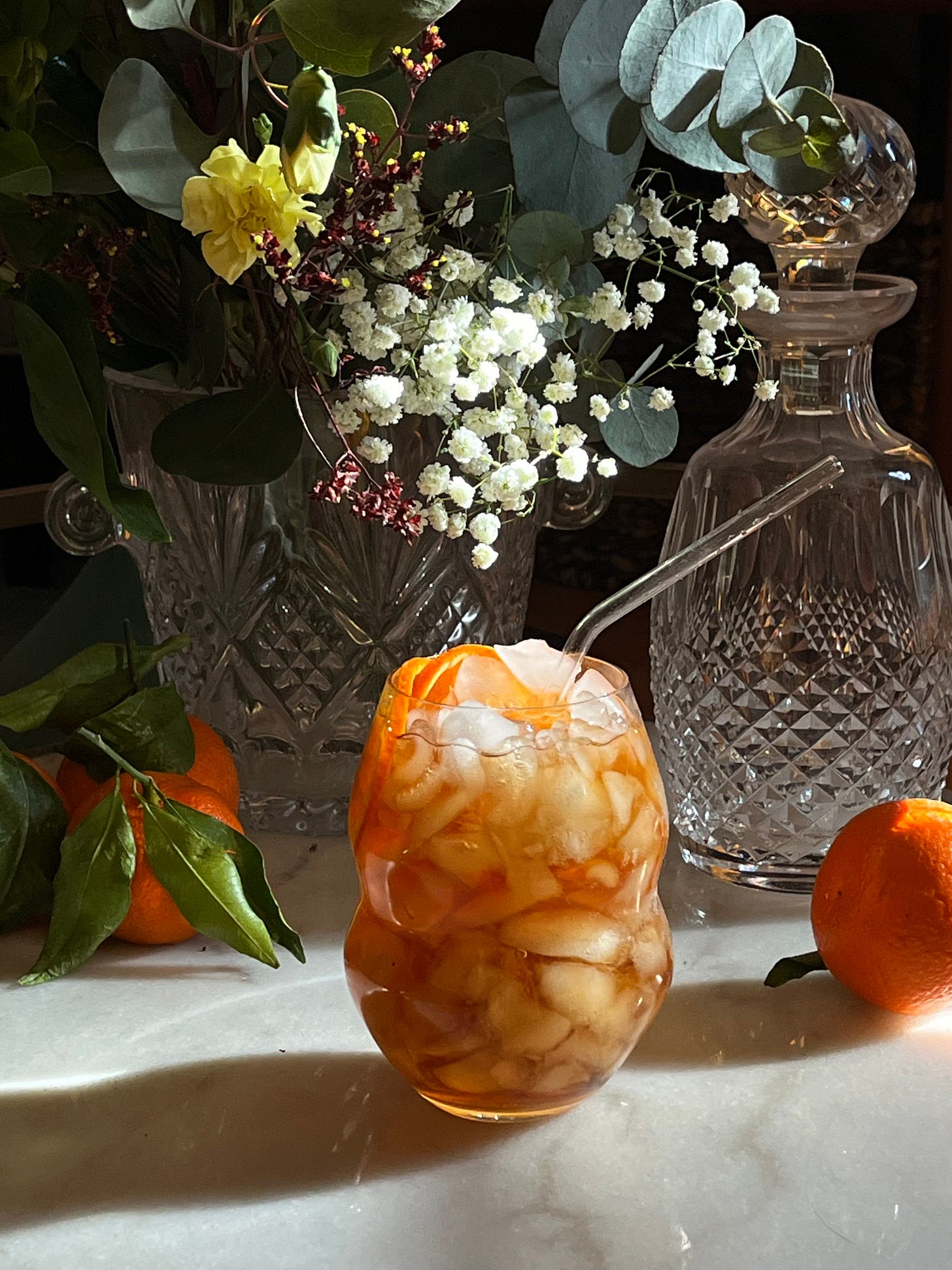 A Sherry Cobbler cocktail served in a wavy glass filled with crushed ice, garnished with orange slices, and a clear straw. The drink is set on a marble countertop with fresh oranges, crystal decanters, and a floral arrangement in the background.