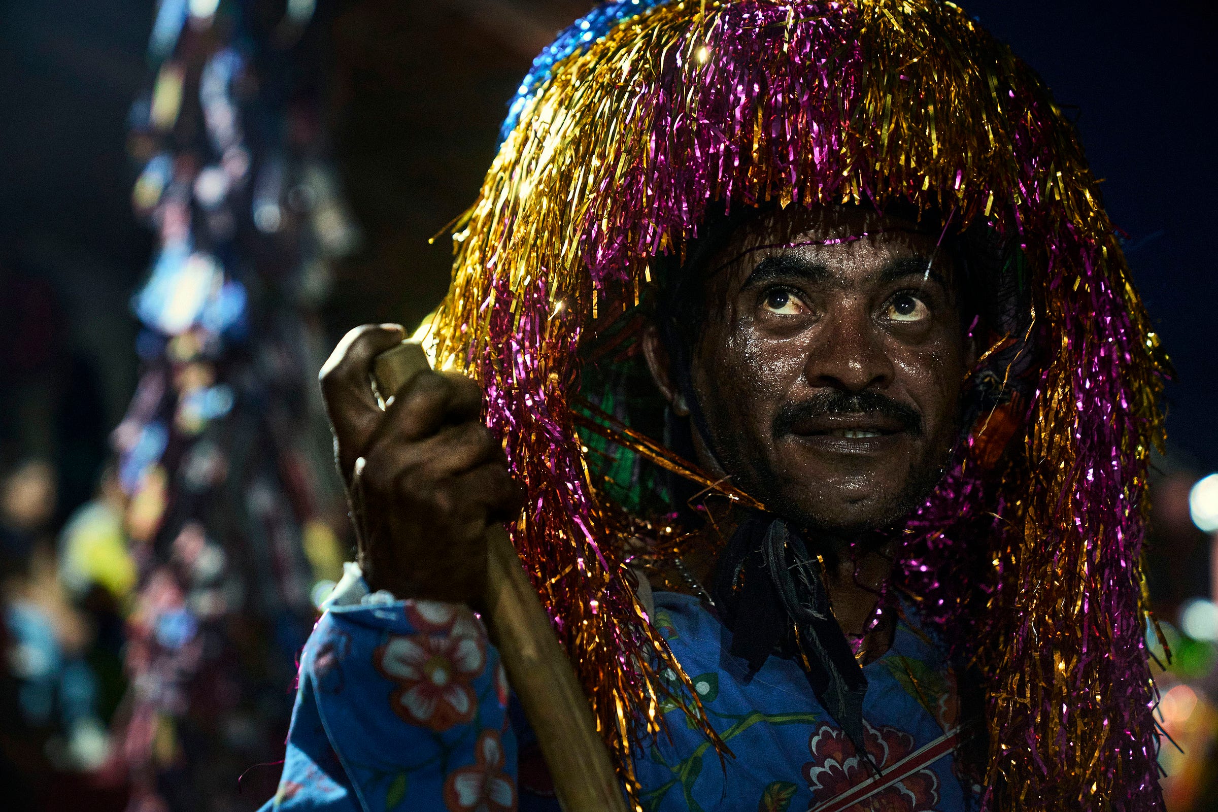 A man in colorful attire holds a stick and wears a vibrant, shiny headdress made of metallic tinsel strands, looking upward. His face is illuminated against a dark background.