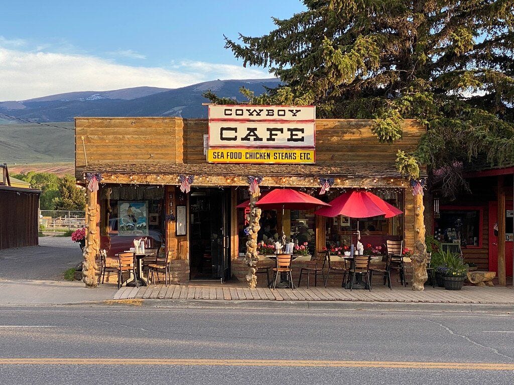 Small-town Wyoming diner with a rustic wooden front, red umbrellas, and mountains in the background.