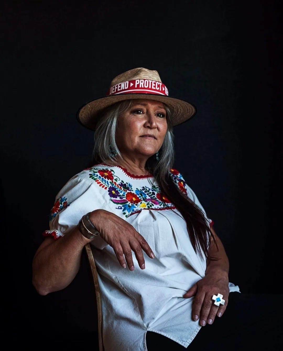 Pennie Opal Plant poses for a portrait. She sits in a chair and wears a hat that reads "Defend, Protect." Pennie Opal Plant poses for a portrait. She sits in a chair and wears a hat that reads "Defend, Protect."
