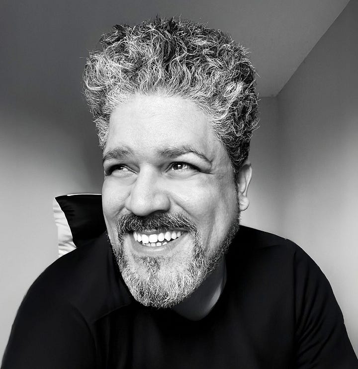 **Alt text:** Black-and-white photo of Christian Ortiz, a smiling person with salt-and-pepper curly hair and a neatly trimmed beard. They are looking slightly to the side, seated indoors in soft natural light, wearing a dark shirt.