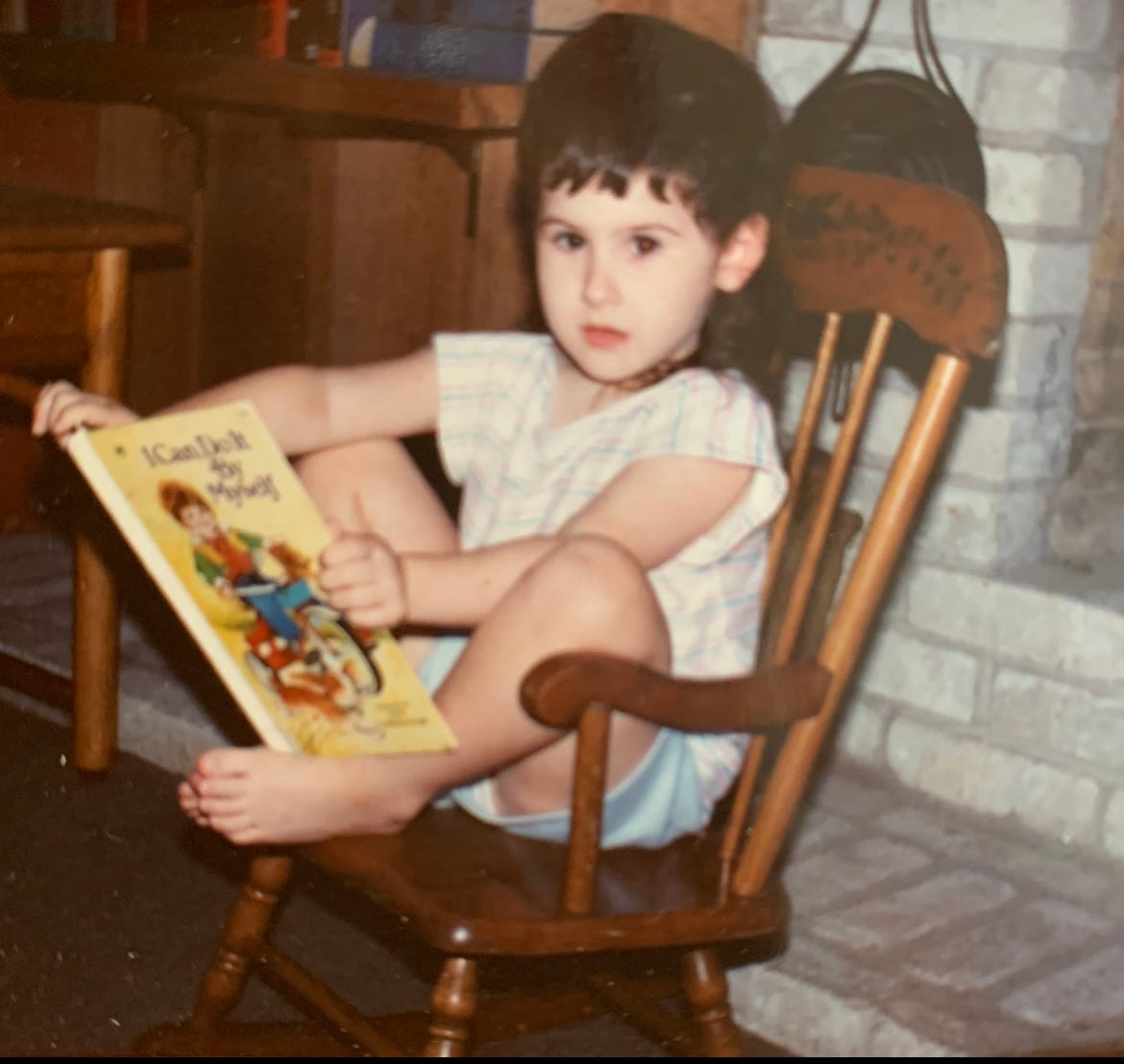 Little Lyric, in a rocking chair, holding a book, “I can do it by myself,” on their feet, looking surprised by the camera. Little Lyric, in a rocking chair, holding a book, “I can do it by myself,” on their feet, looking surprised by the camera.