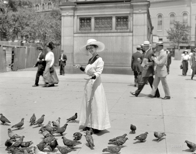 A young woman in a long dress and wide-brimmed hat stands in Boston Common feeding pigeons, smiling slightly as birds gather at her feet, while passersby walk past behind her. Early 20th-century city scene.