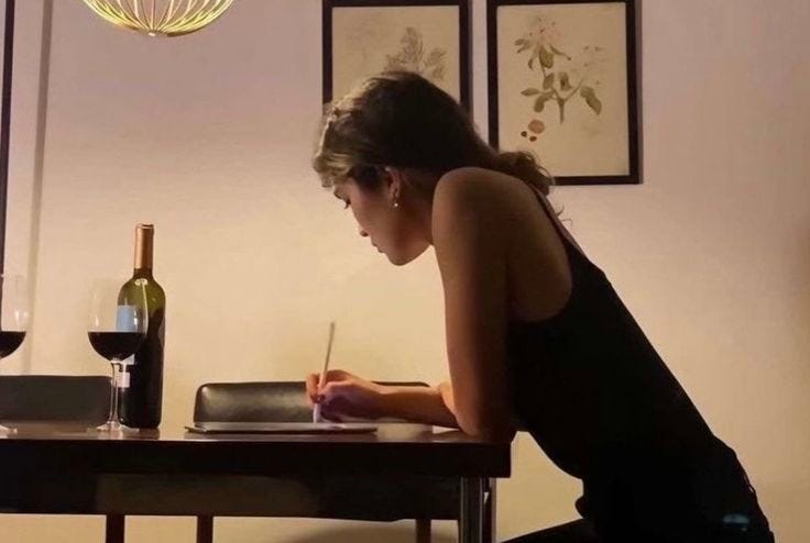This may contain: a woman sitting at a table writing on a piece of paper next to a bottle of wine