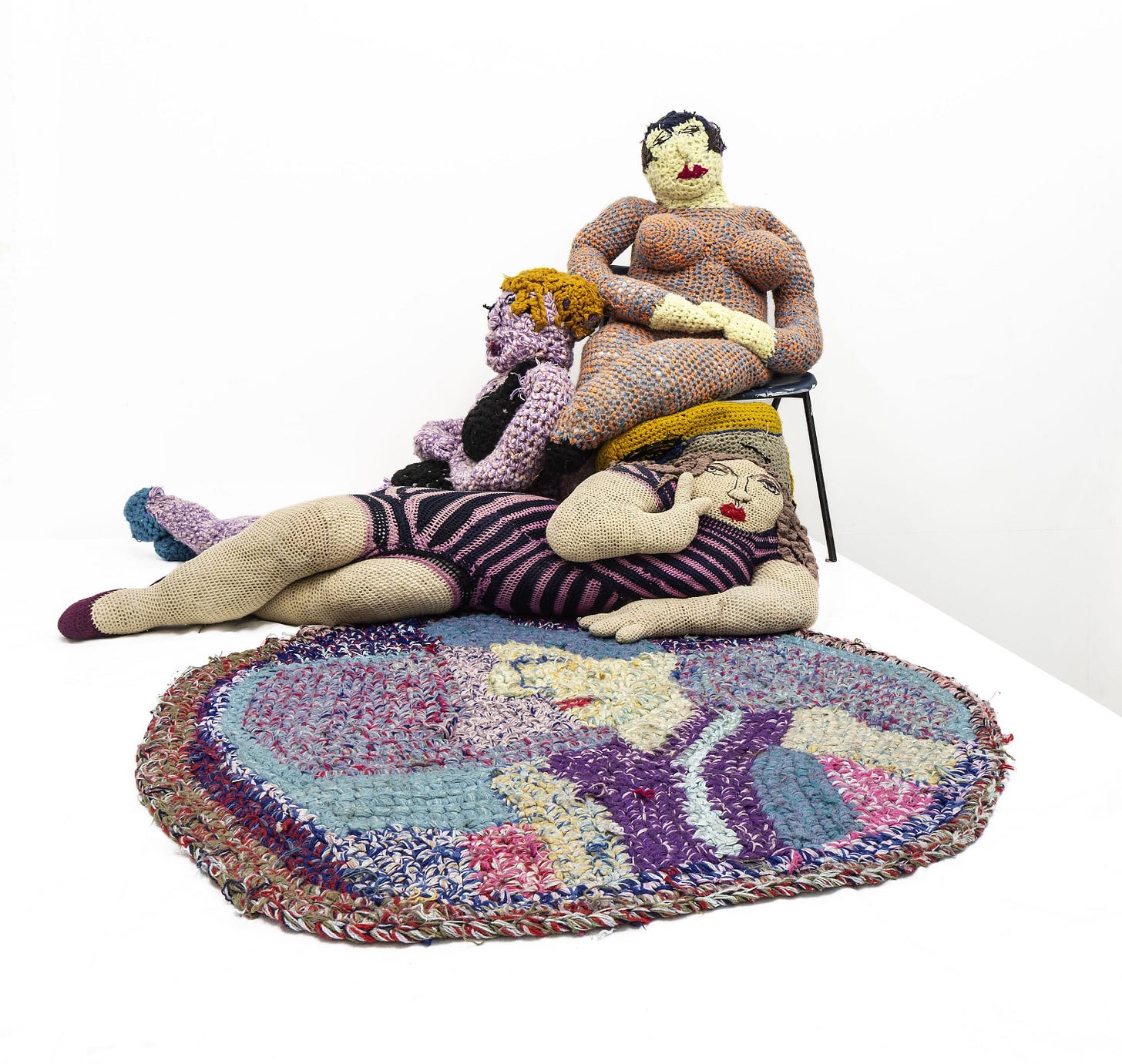 Three life-size crochet women sit together on a rug. One is sitting on a chair, one is sitting up against her on the floor, and the other is reclining on her side. The art work is: Rita McGurn, Untitled Rug and Figures, 1974-1985. Photography by Keith Hunter.