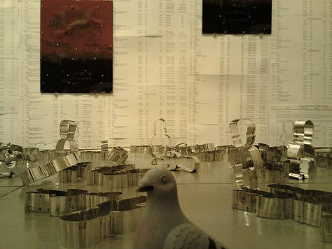A slightly (but artfully) blurry photo that shows several people looking on at Souya Handa and NJCU art department chair in the brightly lit Visual Arts Building Gallery, where the red LED light of one of Handa's pieces can be seen in the background.