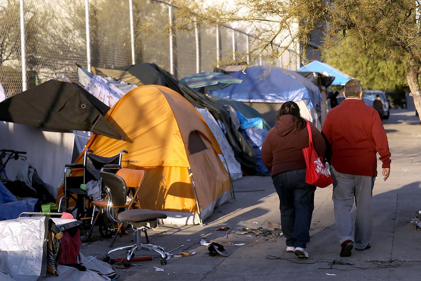 People walking past an assembly of tents in a city's homeless encampment.