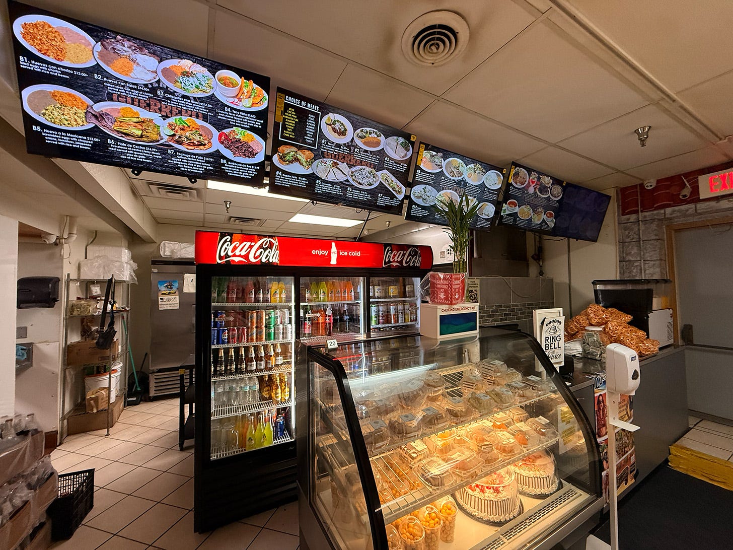 The counter and bakery case with the glowing menu up above. A large Coca-Cola fridge carries an assortment of beverages.