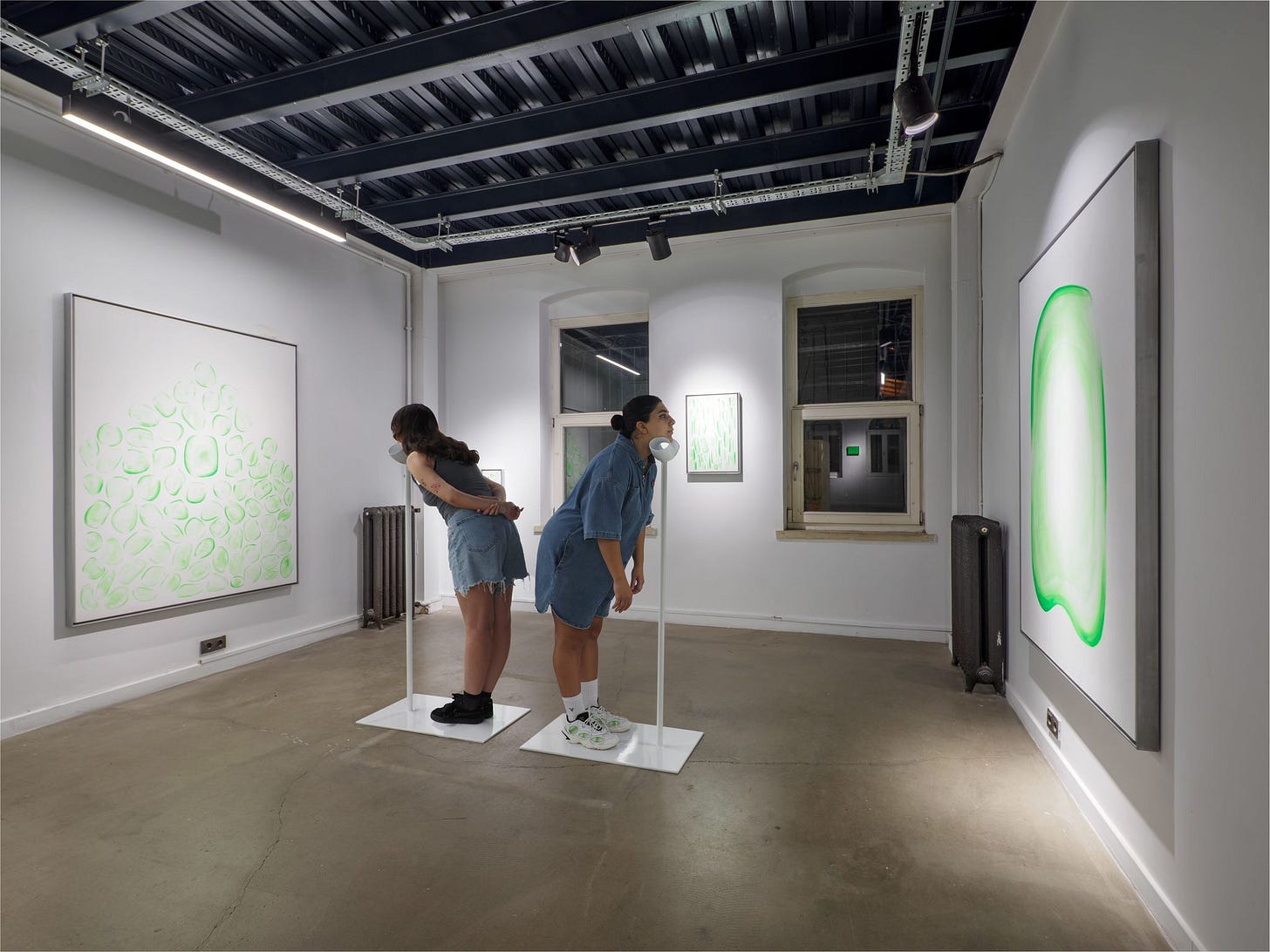 Two people stand and lean into thin white sticks which have chin cups. They appear to view green and white abstract art on the walls of a contemporary art gallery. Two people stand and lean into thin white sticks which have chin cups. They appear to view green and white abstract art on the walls of a contemporary art gallery.