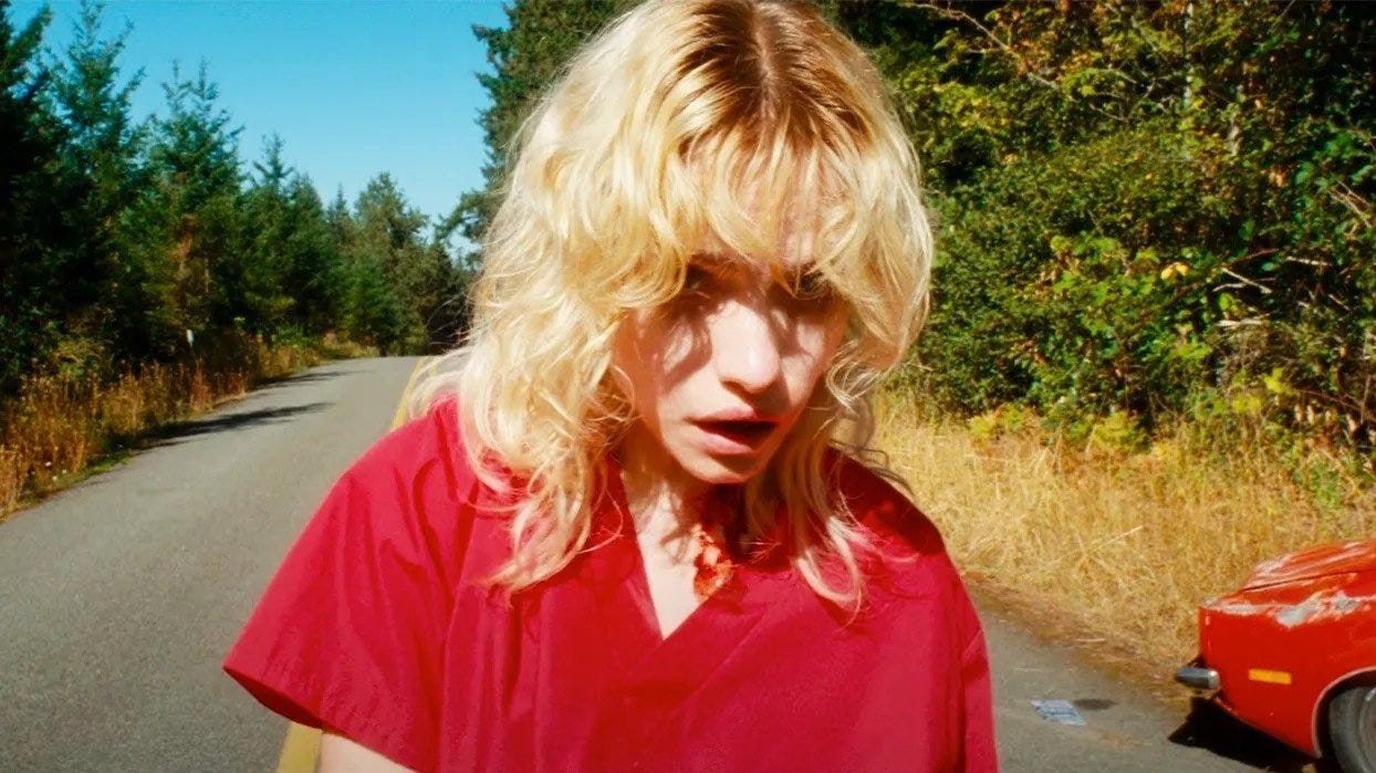 A blonde woman with a bloody neck in a red shirt stands in the middle of a road surrounded by trees.