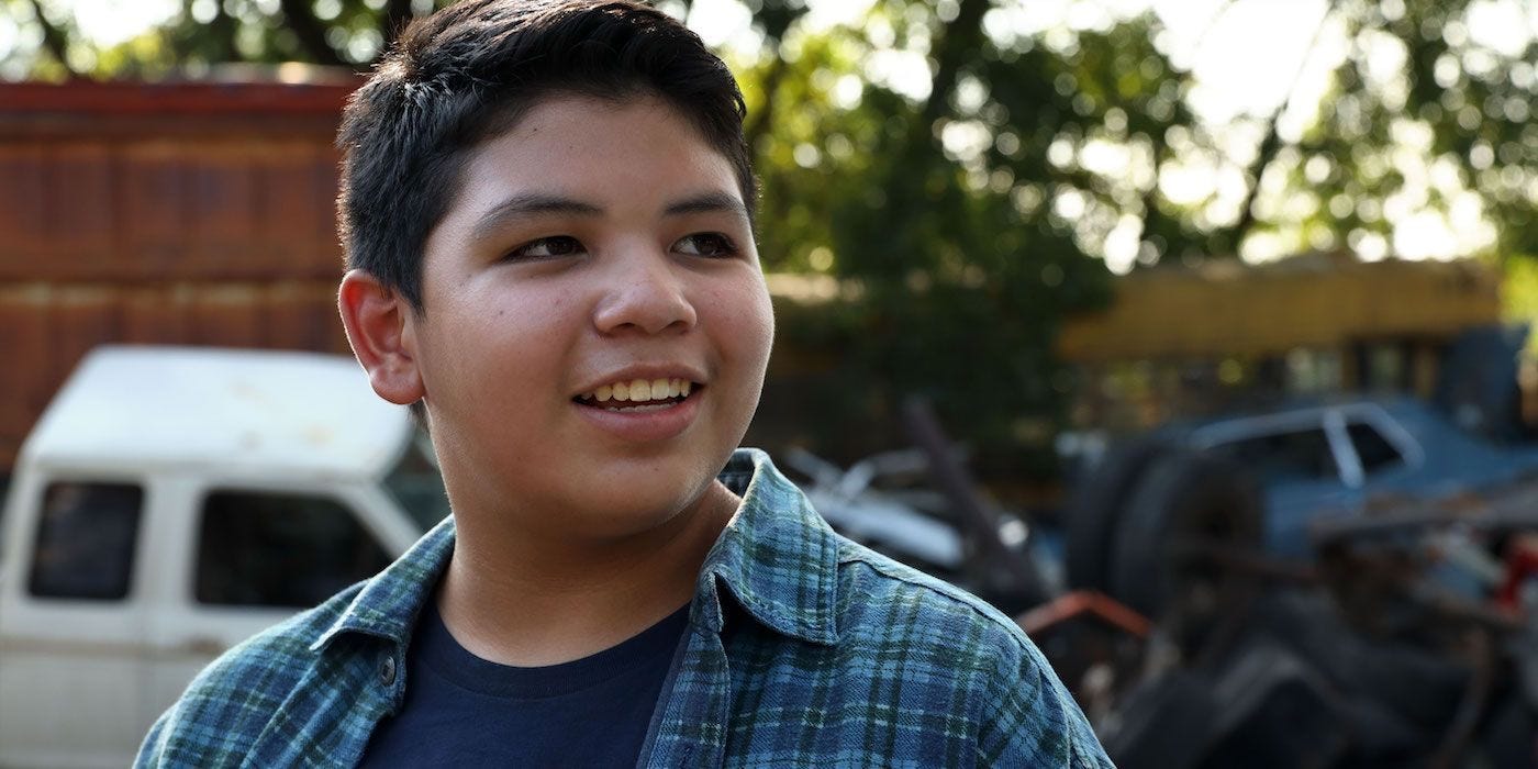 Cheese, a teenage indigenous boy from the show Reservation Dogs, wearing a plaid shirt over a blue T-shirt, looks to the side with a smile on his face.