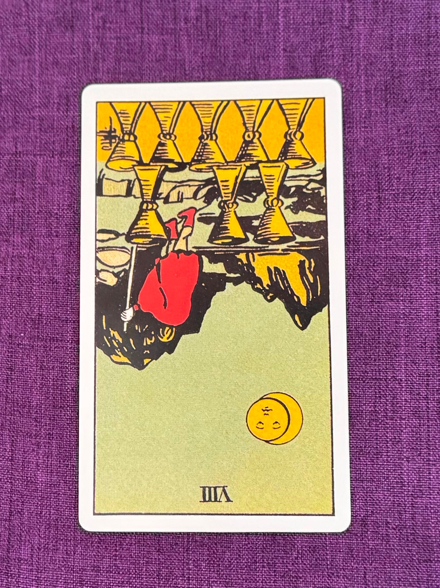 Tarot Card upside down shows a man with a stick walking away in the direction of a mountain. Under his feet are eight golden goblets.
