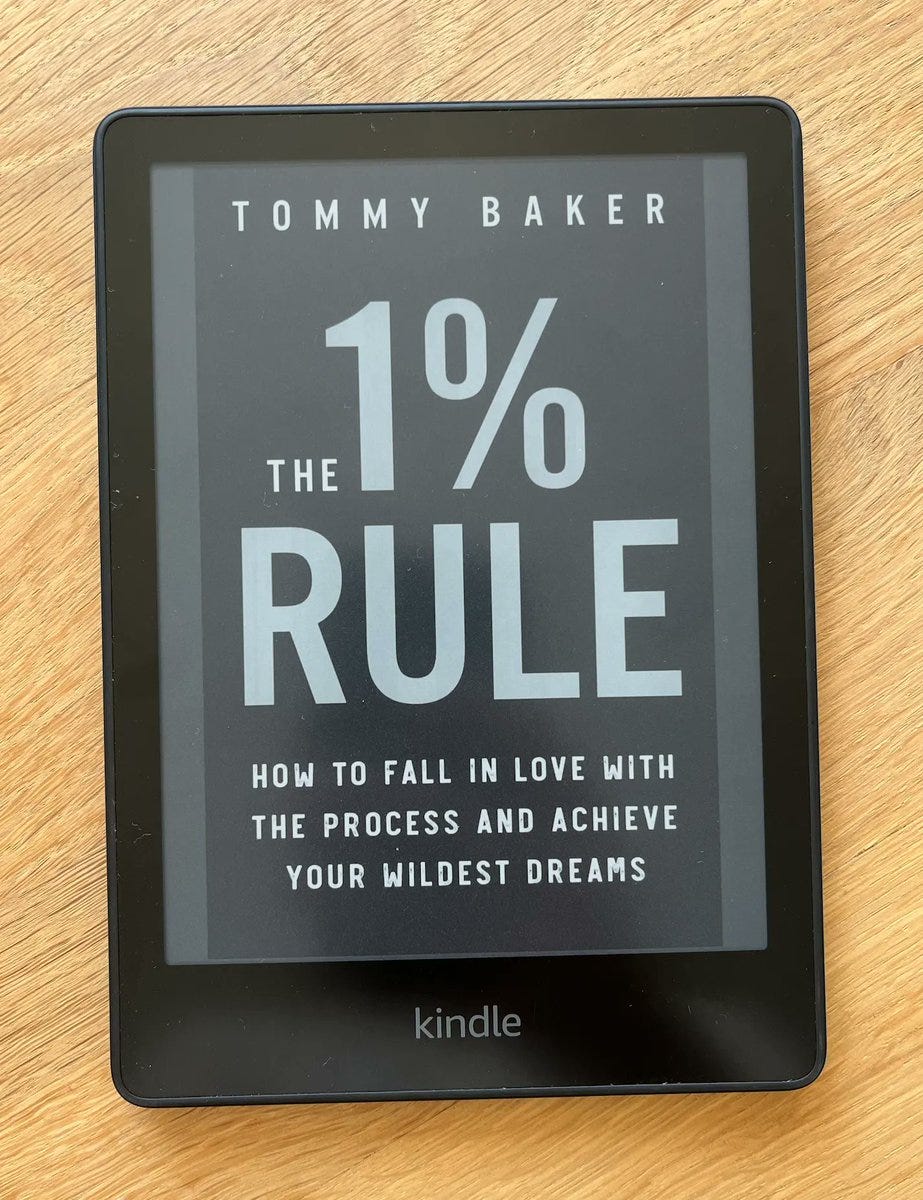 An e-reader displaying the cover of the book "The 1% Rule" by Tommy Baker. The cover features the title prominently in large white text, with the subtitle "How to Fall in Love with the Process and Achieve Your Wildest Dreams" written below it in smaller text. The e-reader is a Kindle, as indicated by the logo at the bottom of the device. The background is a wooden surface.