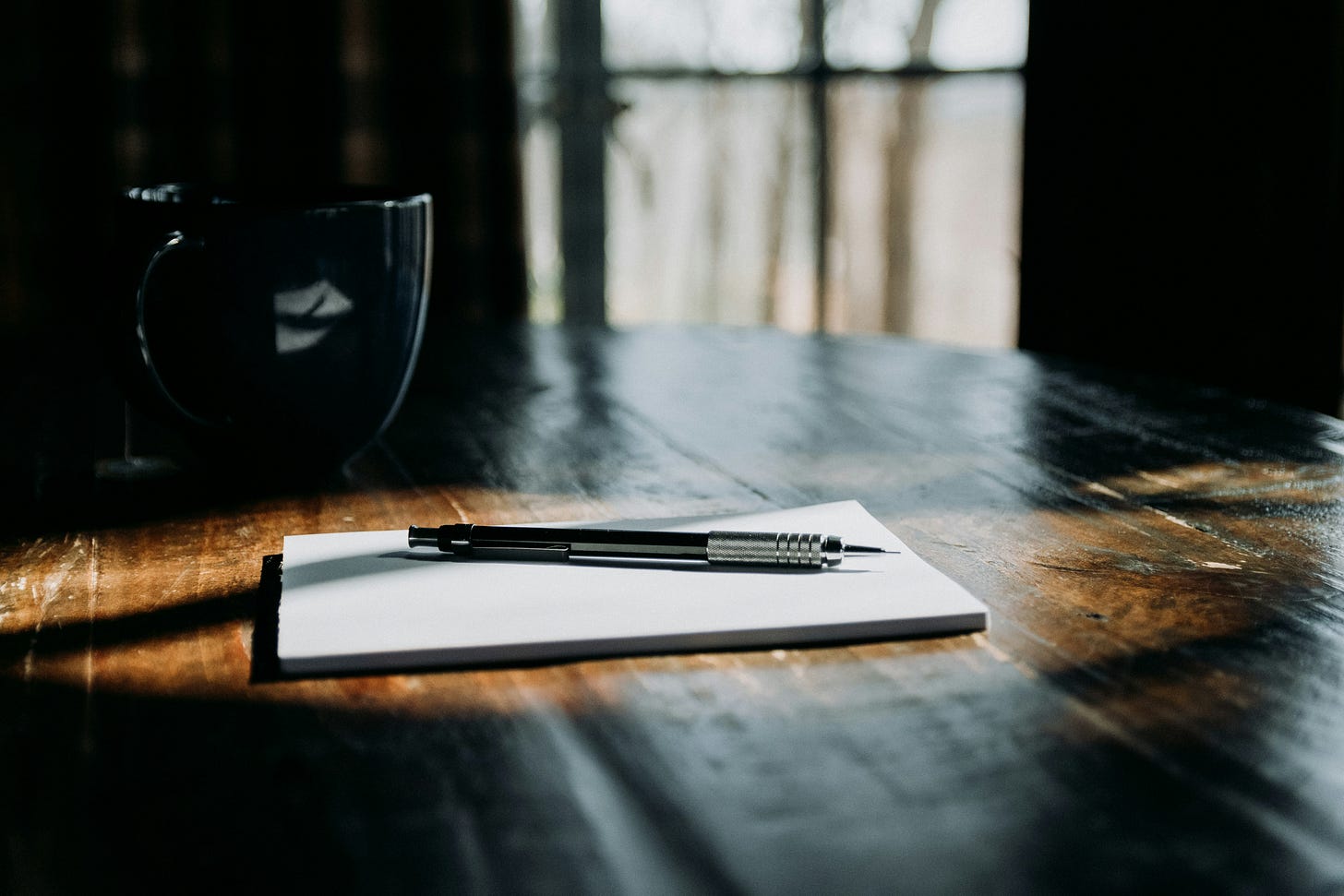 A mechanical pencil rests on a blank white notepad sitting on a dark wooden table. A dark ceramic mug sits in the background near a window with soft natural light filtering through. A mechanical pencil rests on a blank white notepad sitting on a dark wooden table. A dark ceramic mug sits in the background near a window with soft natural light filtering through.