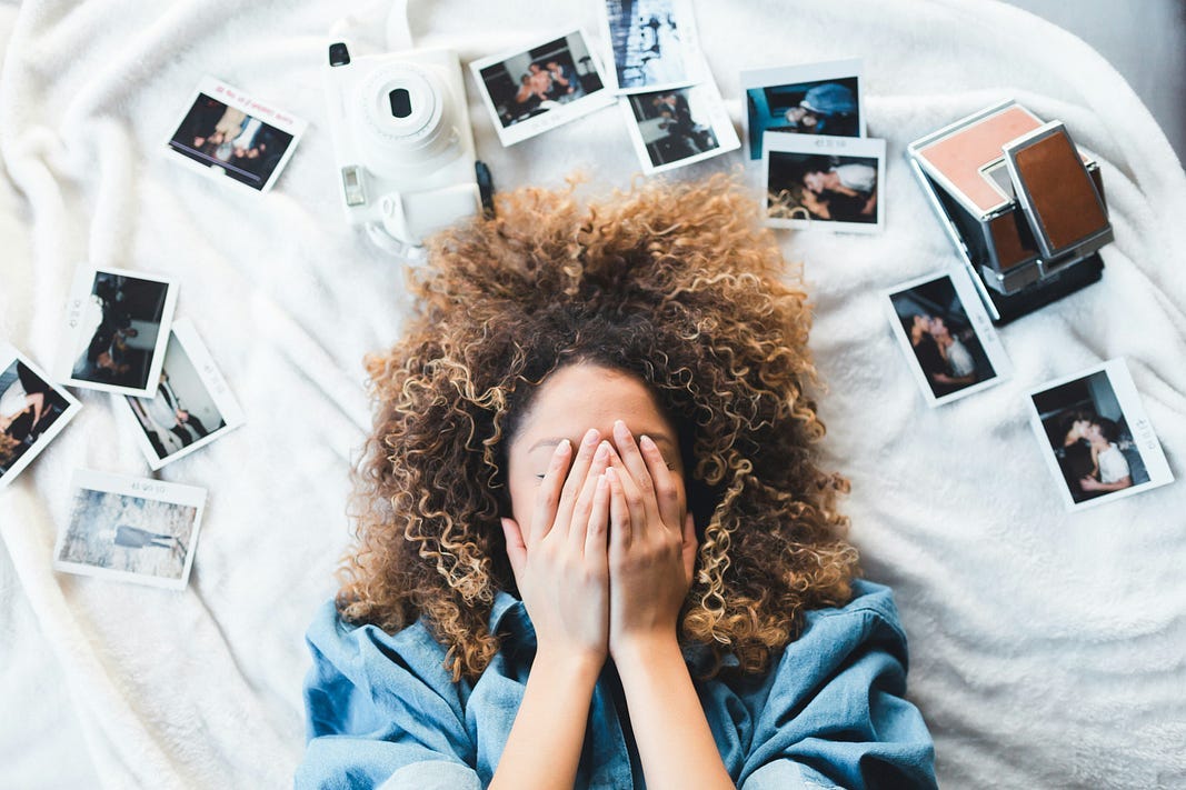 A woman with curly hair lies on a white blanket, covering her face with her hands. Around her are scattered instant photos and two vintage-style cameras, evoking themes of memory, nostalgia, and reflection.