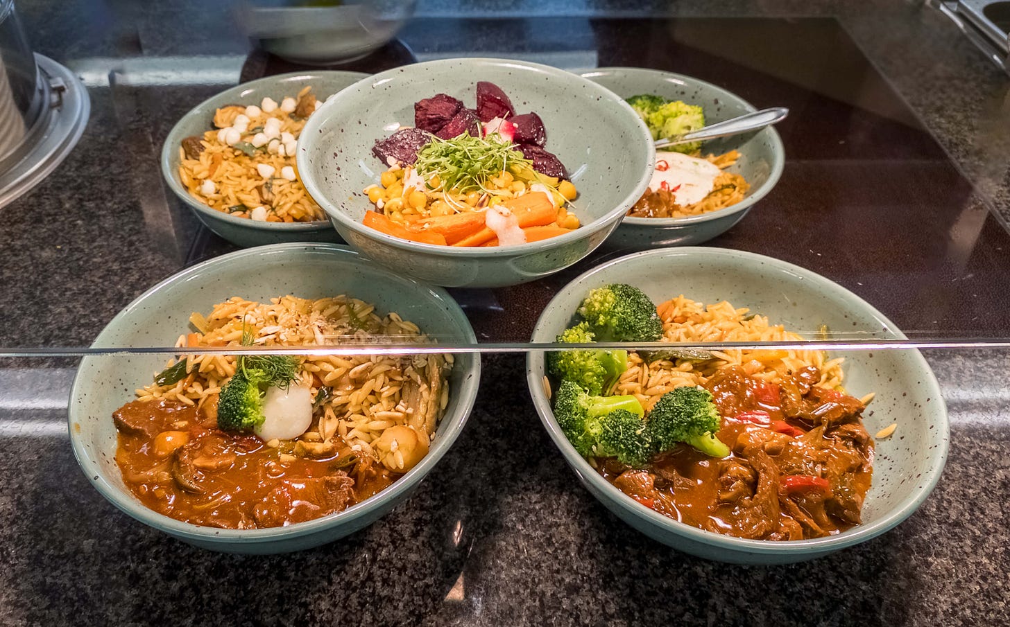 Auslage einer Kantine mit mehreren Keramikschalen, gefüllt mit unterschiedlichen warmen Gerichten. Vorne stehen zwei Schalen mit Orzo, Brokkoli und einer braunen Schmor­sauce mit Gemüse. Dahinter zwei weitere Schalen, eine mit Orzo und knusprigen Toppings, eine mit Orzo, Brokkoli und heller Sauce. In der Mitte eine bunte vegetarische Bowl mit Mais, Rüebli, Randen und Sprossen.