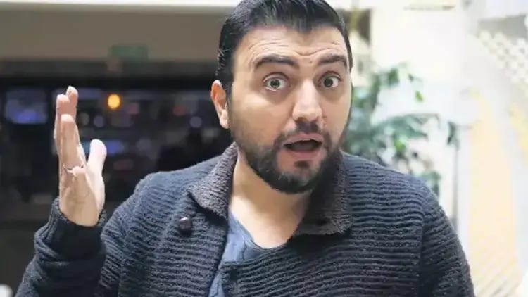 Şaşkın bir ifadeyle elini kaldıran, koyu renkli tekstürlü bir ceket giymiş sakallı Şinasi Yurtsever. İç mekanda çekilmiş bu fotoğrafta, arka planda yumuşak bir aydınlatma görünüyor ve karakteristik abartılı mimikleriyle "Kardeş Payı" dizisindeki rolünü anımsatıyor.