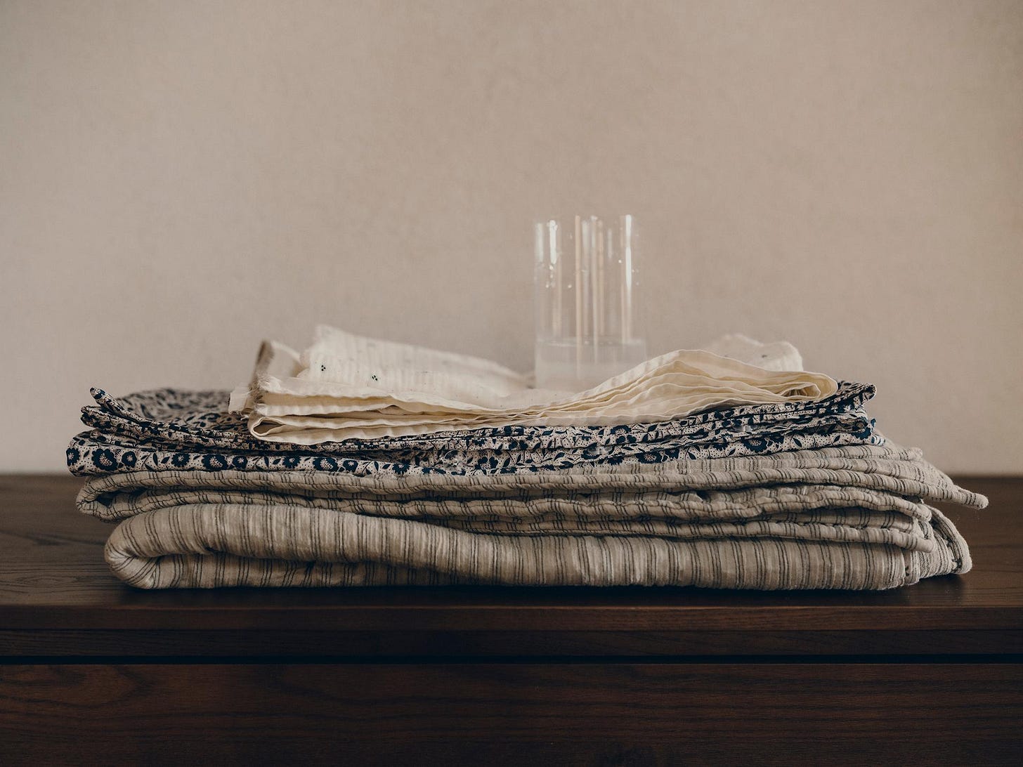 Folded fabric with patterns and textures in neutral tones on a wooden table, with glass tubes in the background.