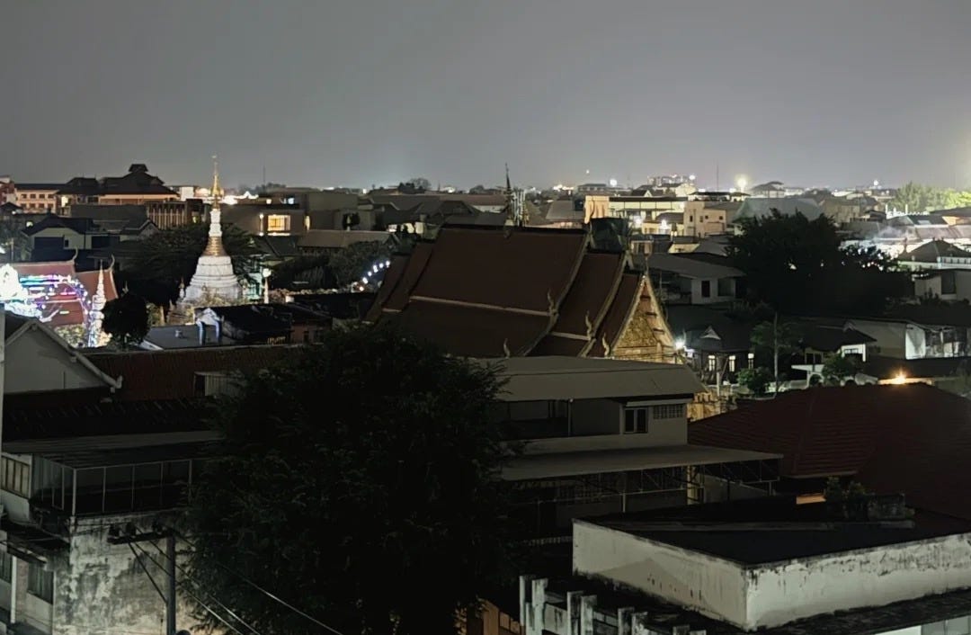 A rooftop view over Chiang Mai at night, looking across the tiered roofs and gilded gable of a Lanna temple and its white chedi to a low-rise skyline of houses and scattered lights.