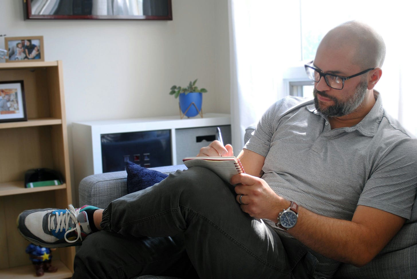Middle-aged man with glasses and beard writing in notebook while sitting on gray couch in home office with bookshelf and plant in background
