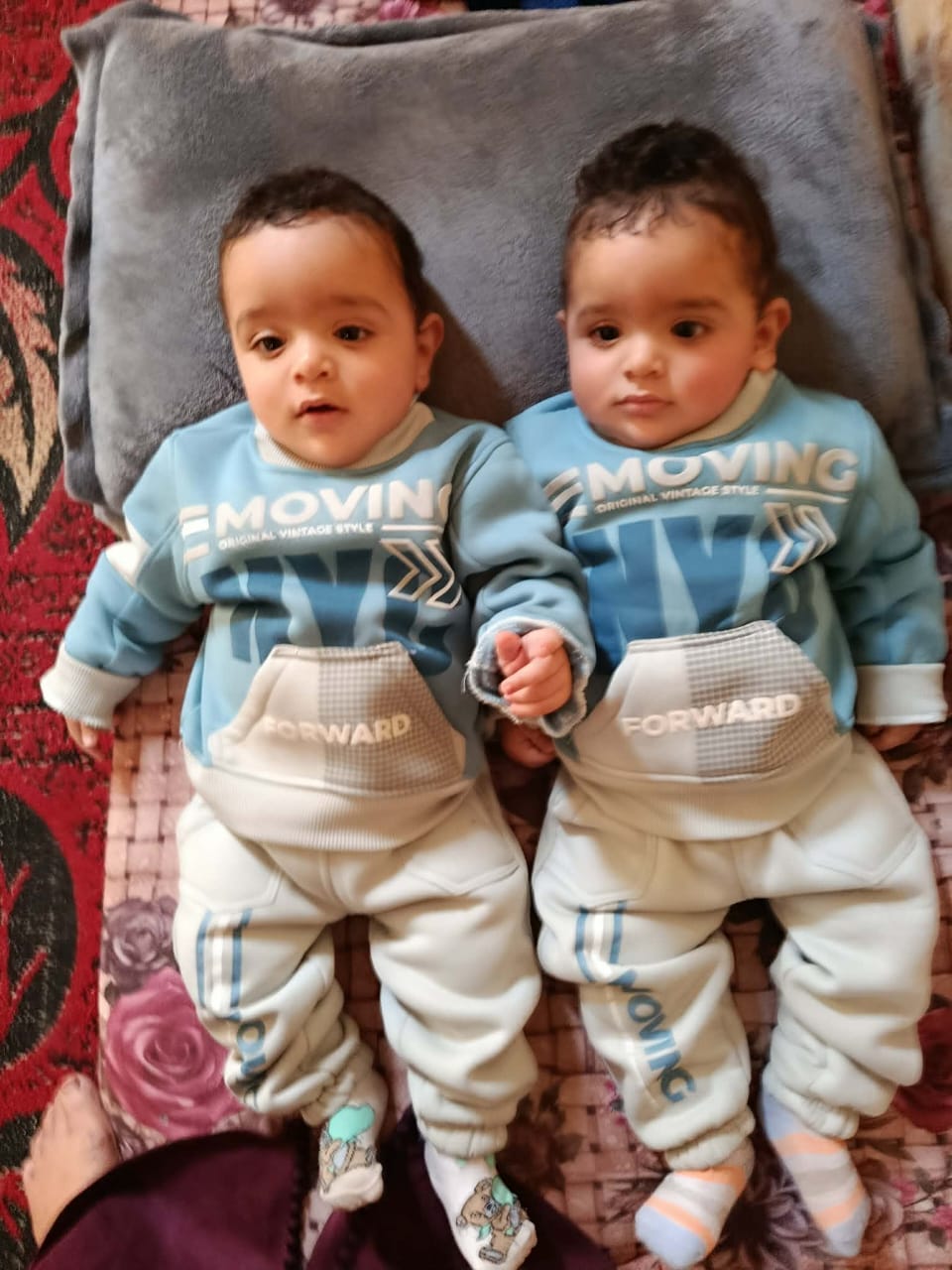Two babies lie side by side on a gray pillow placed on a patterned blanket. They are dressed in matching outfits: light blue sweatshirts with the word “MOVING” printed on the front and cream-colored pants with blue accents and the word “FORWARD” on the pocket. Their heads are close together, and one baby’s hand rests near the other’s hand as they look slightly off to the side. Two babies lie side by side on a gray pillow placed on a patterned blanket. They are dressed in matching outfits: light blue sweatshirts with the word “MOVING” printed on the front and cream-colored pants with blue accents and the word “FORWARD” on the pocket. Their heads are close together, and one baby’s hand rests near the other’s hand as they look slightly off to the side.
