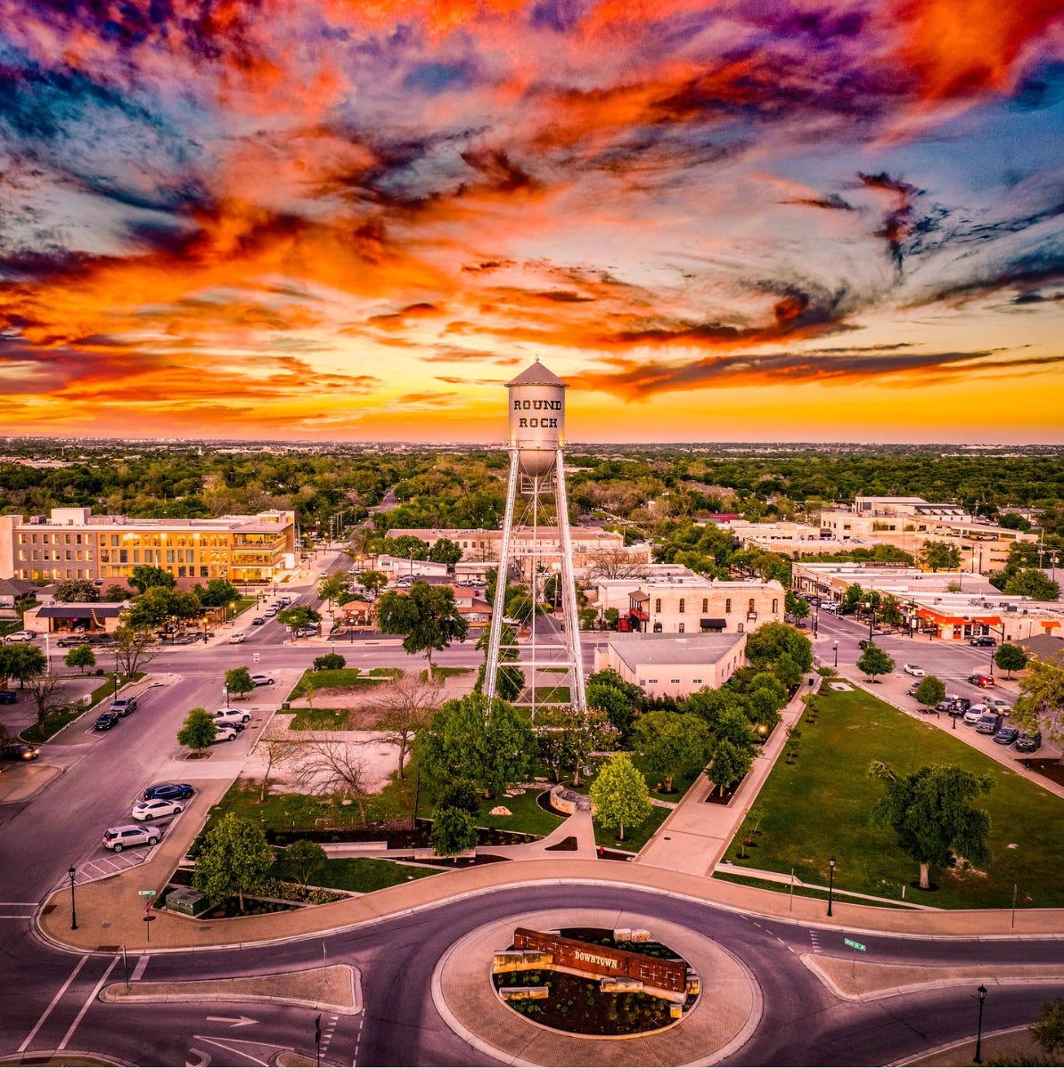 A sunrise aerial view of downtown Round Rock, Texas, featuring the iconic water tower at the center, surrounded by historic buildings, tree-lined streets, and the circular “Downtown” roundabout in the foreground, all under a dramatic, colorful sky.