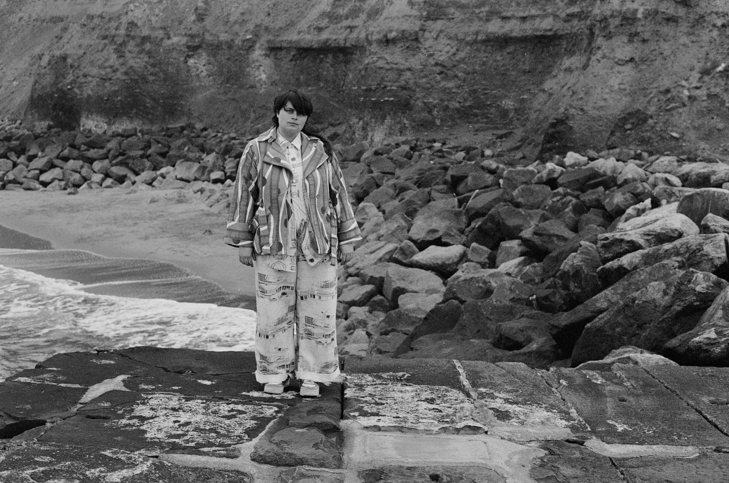 A black and white photo of Laura from Posturing standing on rocks at the coast. A cliff rears up behind her.