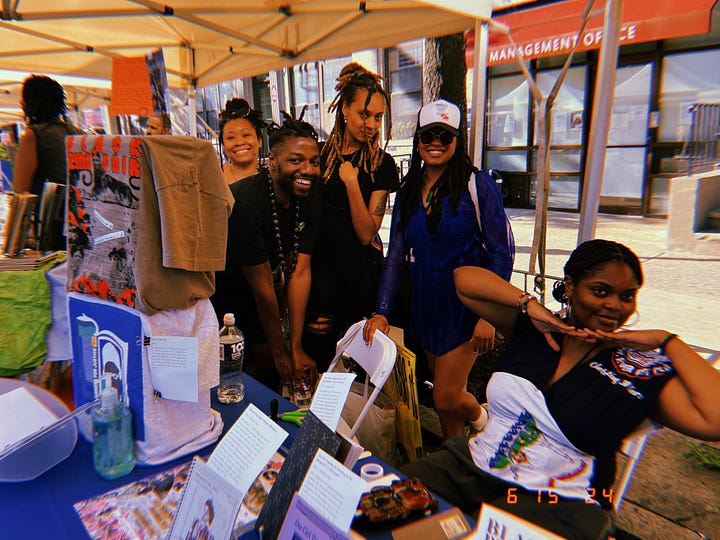 5 Black people standing behind the exhibitor booths smiling and posing at the festival.