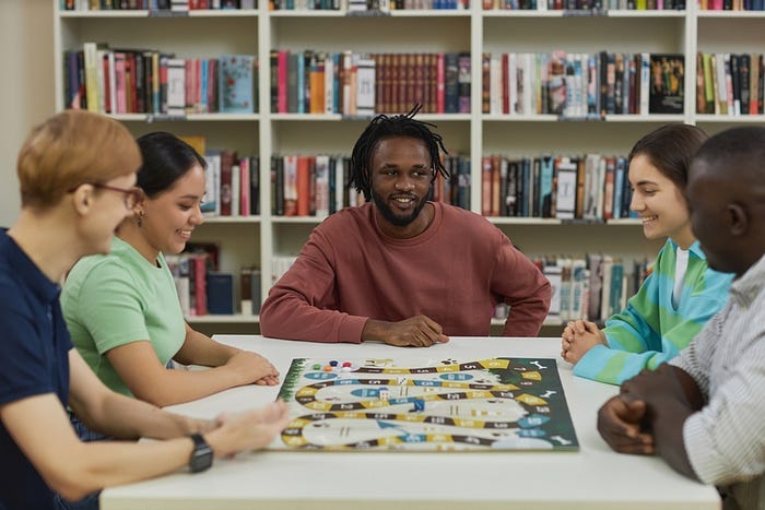 #descrevemospravocê  Cinco pessoas estão reunidas ao redor de uma mesa branca, jogando um jogo de tabuleiro em um ambiente que parece ser uma biblioteca ou sala de leitura. Ao fundo, há uma grande estante repleta de livros organizados em fileiras verticais e horizontais, coloridos e de diferentes tamanhos. O grupo é diverso, com homens e mulheres de diferentes tons de pele, cabelos e estilos. Estão todos sorrindo ou demonstrando concentração, interagindo de maneira descontraída. No centro da imagem, um homem com cabelos crespos e amarrados em pequenas tranças usa uma blusa de manga comprida em tom terroso e observa o tabuleiro com atenção. À sua esquerda, uma mulher de cabelos escuros presos e camiseta verde-clara também olha para o jogo, sorrindo. À esquerda dela, outro homem de cabelos ruivos curtos e óculos, com camiseta bege e colete azul, sorri enquanto segura uma das peças do jogo. À direita da imagem, uma mulher de cabelos castanhos escuros, lisos e soltos, veste uma blusa de listras verdes e azuis e observa o tabuleiro com um leve sorriso. Ao lado dela, um homem de pele escura e cabelo raspado, usando camisa branca com listras finas, parece conversar com o grupo. O tabuleiro é colorido, com caminhos curvos, casas numeradas e pequenas peças de jogo em diferentes cores, como vermelho, azul e preto. A atmosfera é acolhedora, com foco em convivência e lazer coletivo.
