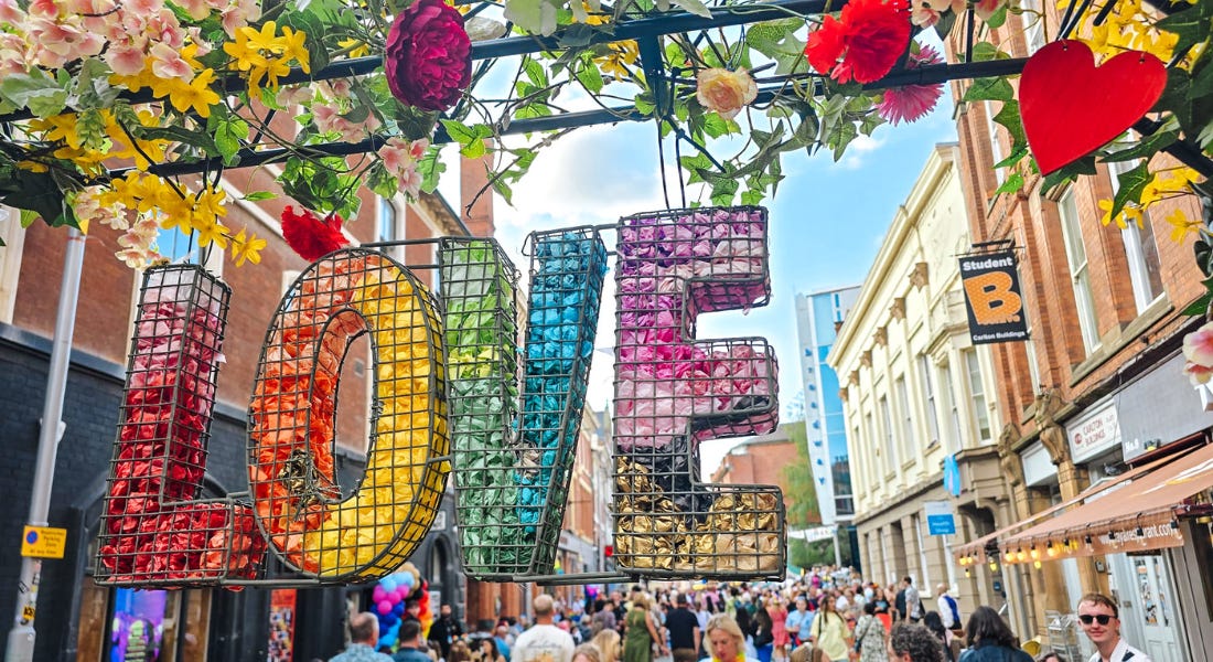A 'Love' sign with rainbow coloured flowers in it with a flower arch above it and people below it A 'Love' sign with rainbow coloured flowers in it with a flower arch above it and people below it