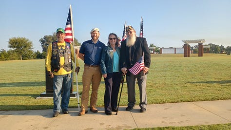 A group of veterans and community members pose outdoors at a POW*MIA memorial. In the foreground is a black granite monument engraved with the POW*MIA emblem and the words “YOU ARE NOT FORGOTTEN,” with a small American flag at its base. Several U.S. flags stand behind the group. Some people wear military dress uniforms and VFW-style caps; one cap reads “Tallahassee,” and another hat bears the U.S. Air Force insignia. On the right, an elderly veteran in a wheelchair salutes while a man in a suit speaks with him. The scene is on a sunny day in a grassy field with cars and flagpoles in the background.