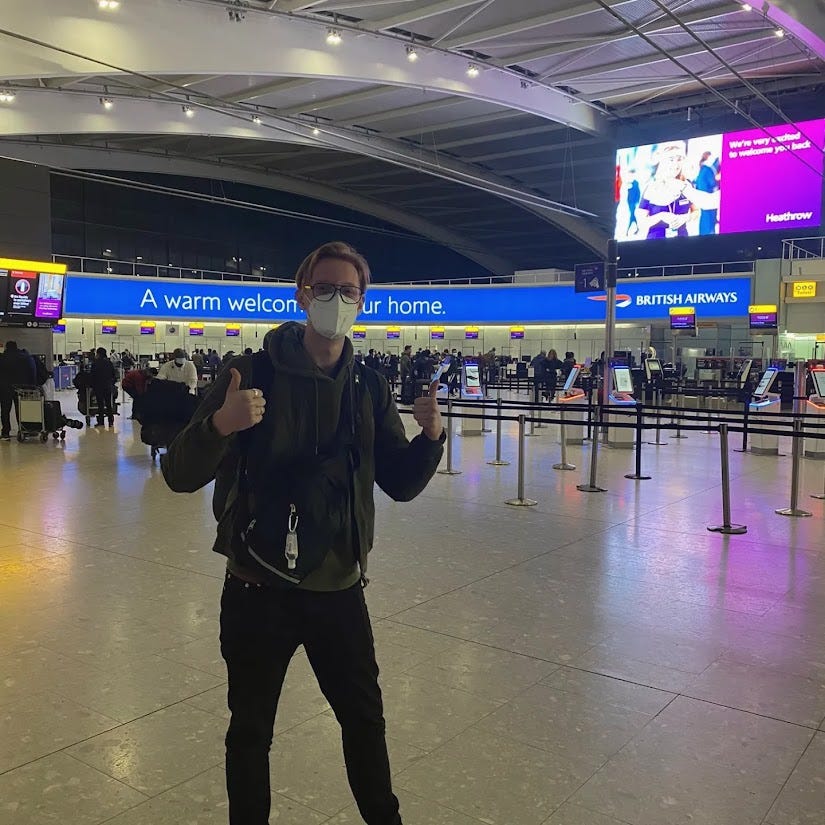 Me at the airport, giving a thumbs up with very little luggage and a face mask on