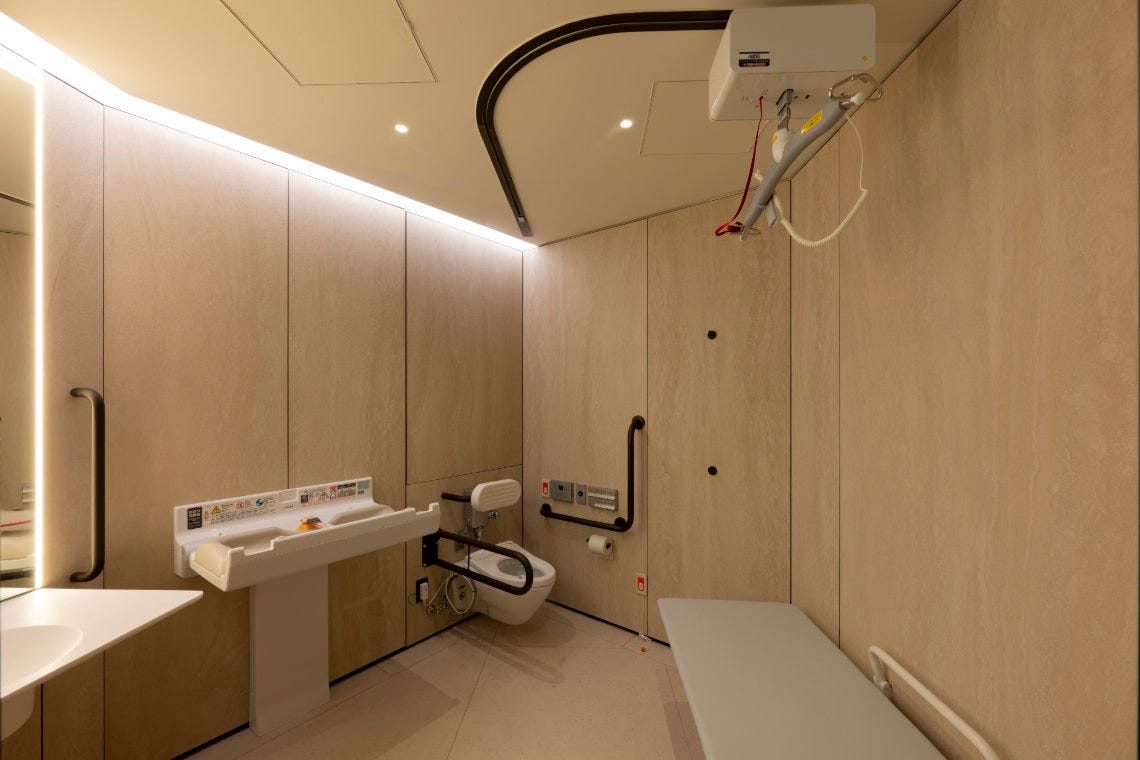 Spacious accessible restroom with a ceiling lift, wall-mounted toilet, baby changing table, and padded bench.