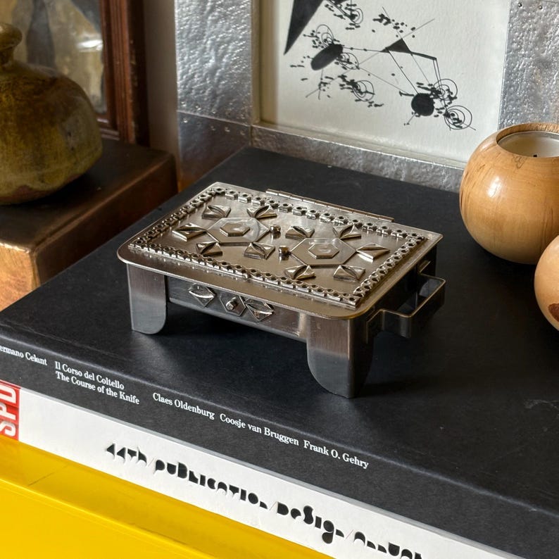 May include: A small, silver-toned metal box with a hinged lid. The lid and sides are decorated with geometric patterns. The box has short, rectangular legs and a handle on one side. It sits on a stack of books.