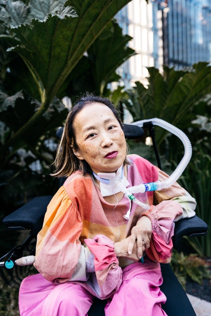Alice Wong, Asian American woman in a wheelchair with a tracheostomy at her neck connected to a ventilator. She’s wearing a pink plaid shirt, pink pants, and a magenta lip color. She is smiling and behind her are a bunch of tall prehistoric looking plants. Photo credit: Allison Busch Photography.