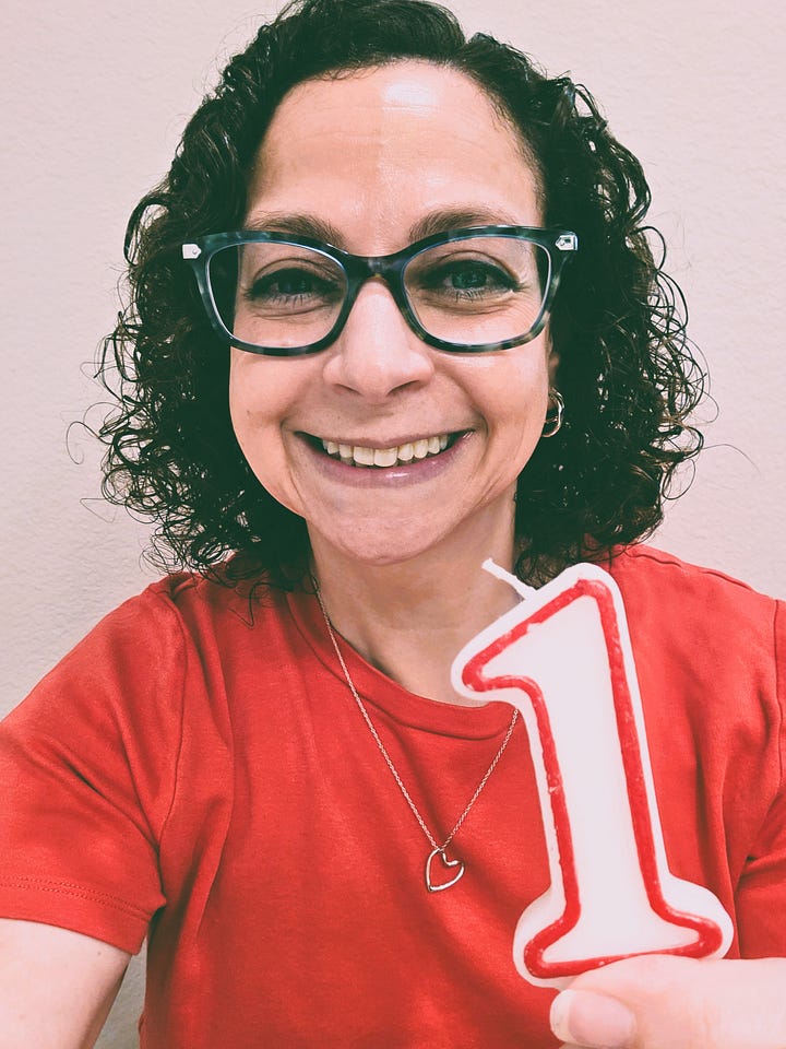 Photo 1: A photograph of the writer of this article, Debra Rafson. She has curly, brown, shoulder-length hair. She is wearing glasses, a red t-shirt, and a heart-shaped silver necklace. She is holding up a birthday candle shaped like the number “1,” in celebration of one year writing “What’s Her Problem?” on Substack. Photo 2: A photograph of Debra Rafson on her actual 1st birthday. She is wearing a pink dress, white tights, and black shoes. She is sitting on a pink blanket, which also contains a birthday crown and a “1” birthday candle. She is holding up a party hat in her right hand, and there are 2 balloons stuck to the wall behind her.