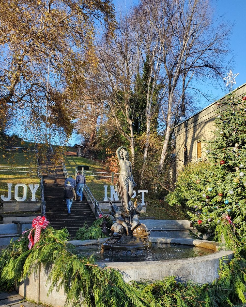 Color photograph of the Haller Fountain and Taylor Street stairs in Port Townsend decorated for the holiday season. Greenery, wreaths, and large white letters spelling “JOY” appear around the stairs, with the historic flagpole visible near the top right.