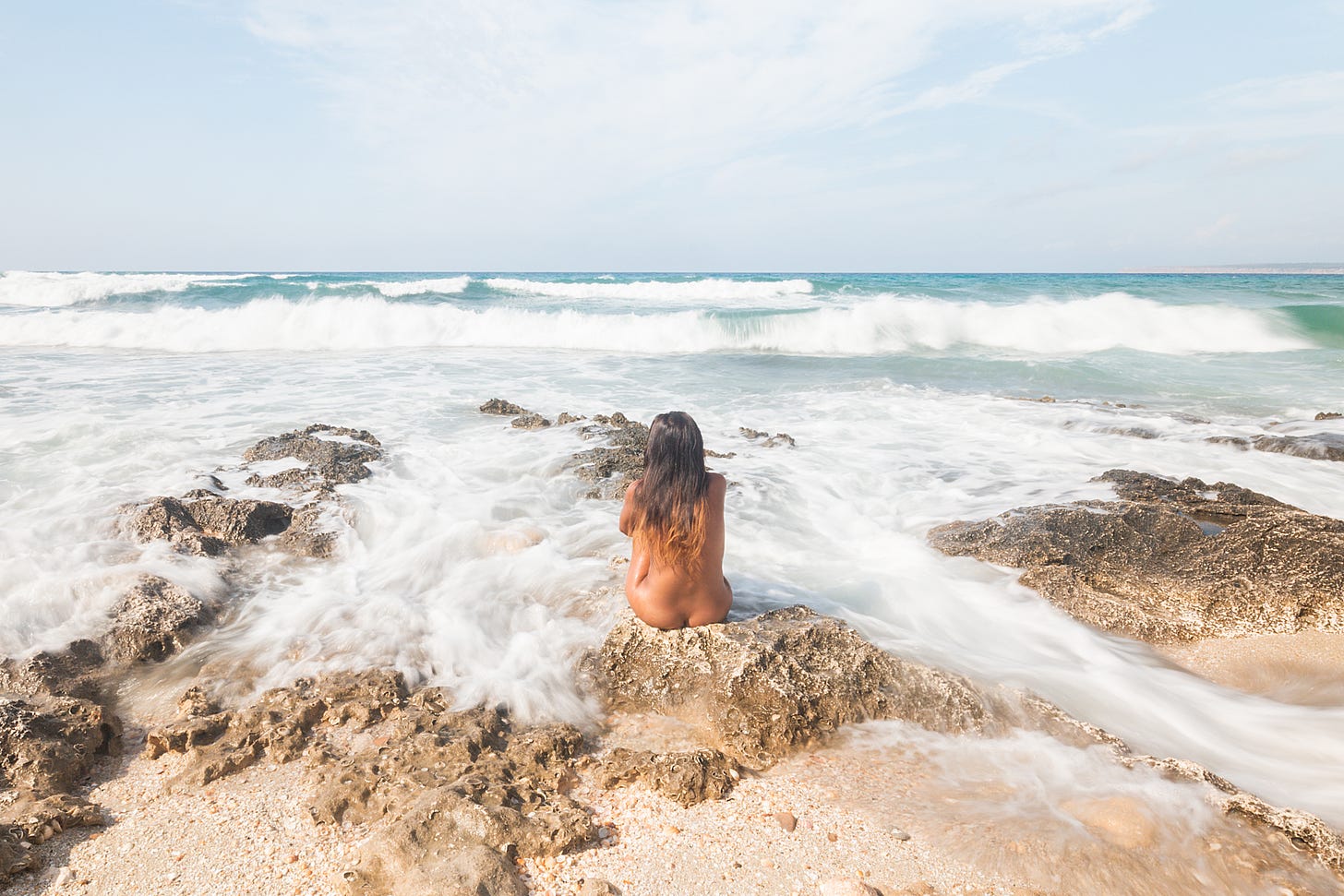 Nude woman sits on a rocky shore facing the ocean. 