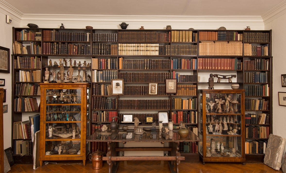 “A cozy room with a large, dark-wood bookshelf filled with neatly arranged old books and artifacts. Two glass display cabinets, one on the left and one on the right, contain small sculptures and figurines. A wooden table in front of the bookshelf holds pottery and other artifacts. The room has a warm, vintage feel with parquet flooring and framed art on the walls.