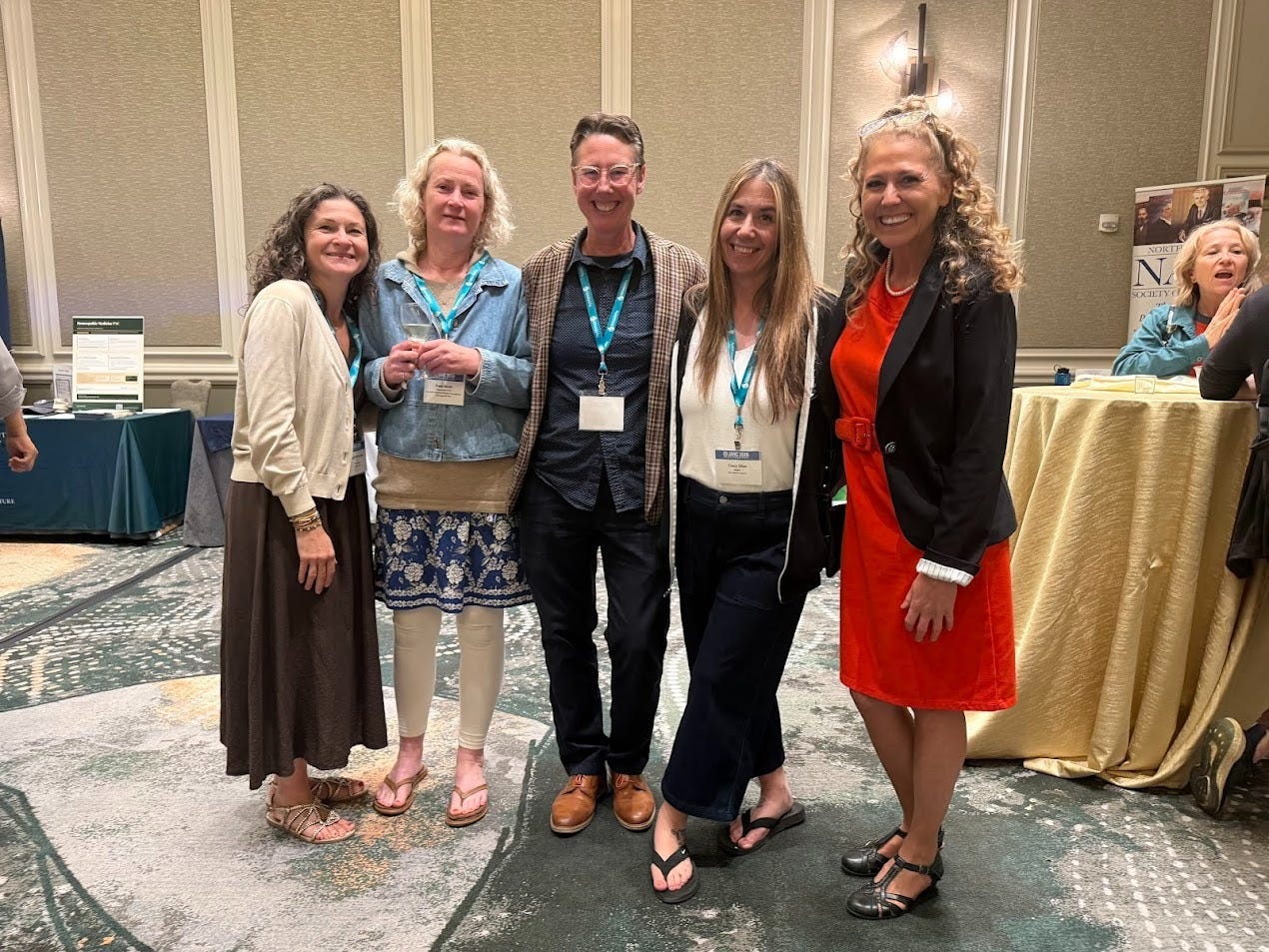 Five women in business-casual attire smile together wearing teal conference lanyards at the Joint American Homeopathic Conference. 