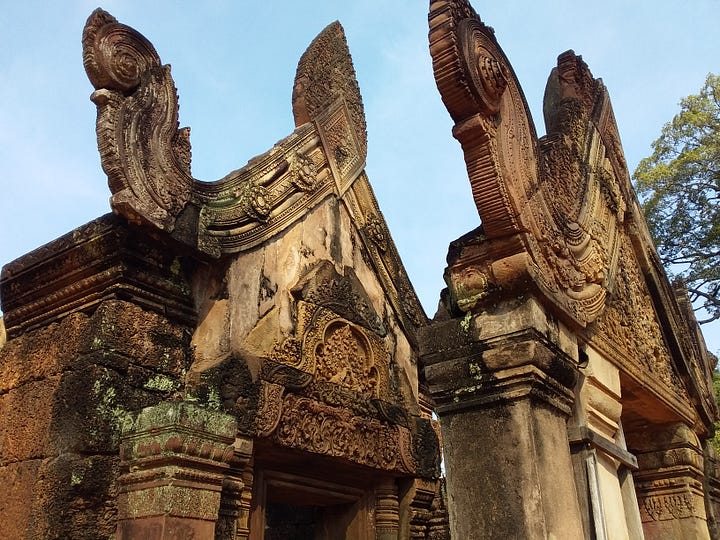 A side-by-side image. The left photo shows a close-up of highly intricate, ornate carvings on the reddish-brown stone roof and pediment of an ancient temple. The right photo shows a wider view of a multi-tiered temple tower made of the same detailed reddish stone, set against a bright blue sky and surrounded by green trees.