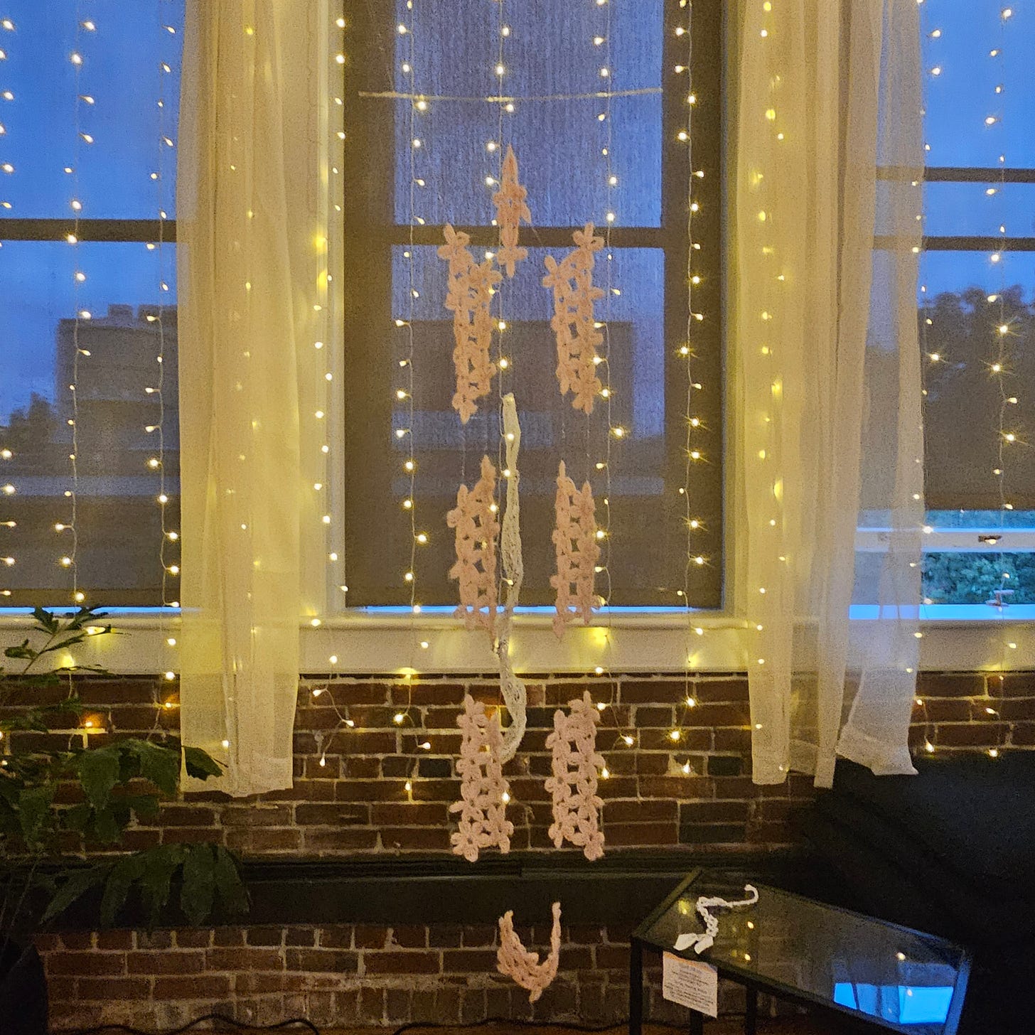  Pretty Pain hanging from the ceiling. It has invisible thread holding pink embroidered pieces together in front of a window illuminated with small fairy light